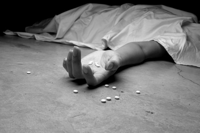 A black and white image of an addict, dead beneath a sheet, arm outstretched, pills in the palm of their lifeless hand and littering the floor near their hand.