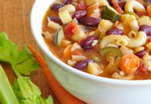 Minestrone Soup A bowl of minestrone soup with two pieces of celerey and one carrot on the table next to it.