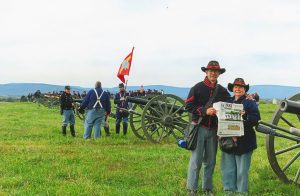 The Dazeys stand dressed in Union soldier uniforms on the battle field.  A group of Union soldiers stand behind them near an Civil War era cannon.