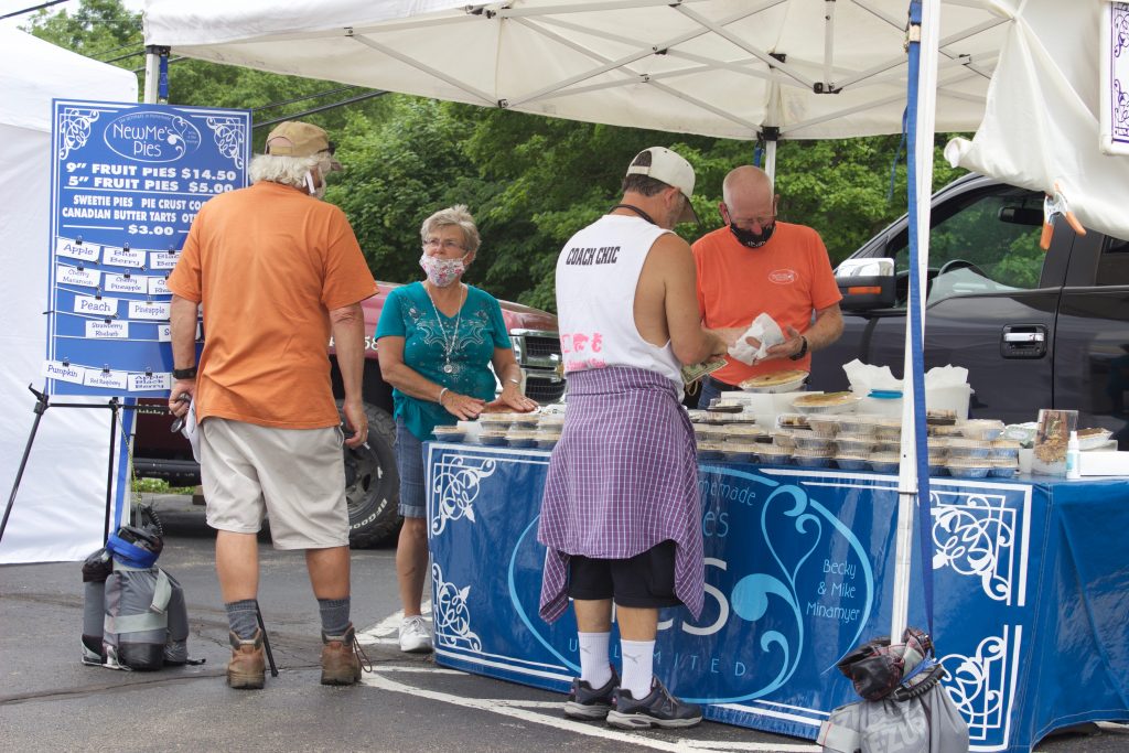 A couple talks to two customers at their pie stand at a farmers market.