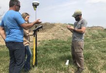 Three people stand in a field with GPS surveying equipment.