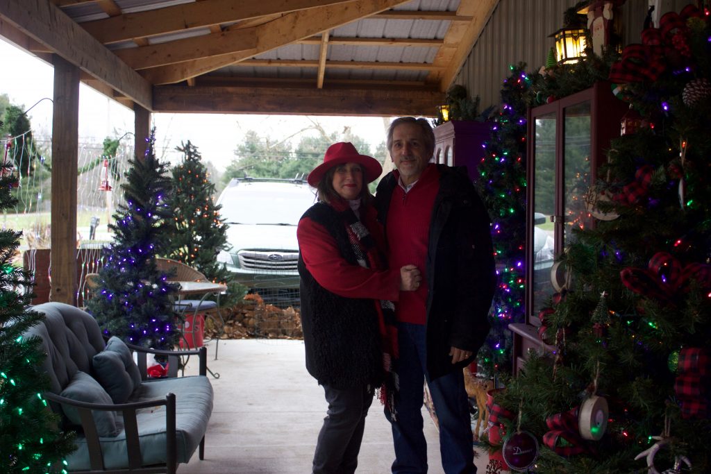 A woman and a man stand outside a pole building decorated with Christmas lights and trees.