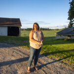 Balancing love, loss and life after a farmer’s suicide A woman wearing jeans and a light-colored top stands on a gravel driveway, framed in the background by a small open barn and a tree. Behind her are soybeans in a field.