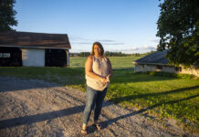 Balancing love, loss and life after a farmer’s suicide A woman wearing jeans and a light-colored top stands on a gravel driveway, framed in the background by a small open barn and a tree. Behind her are soybeans in a field.