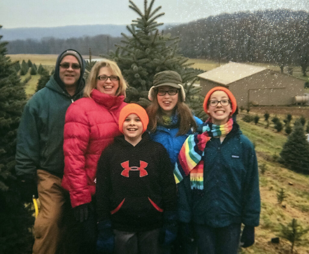 A father, mother, two preteen daughters and their son stand in a field of pine trees. They're all looking at the camera and smiling.