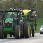Farmers ask drivers to be patient during spring planting season A tractor pulling a planter drives on a road with a car following closely behind it.