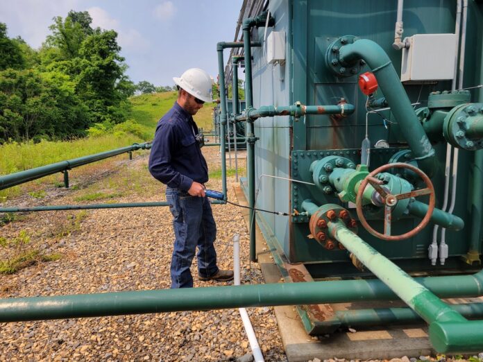 A worker holds a handheld gas detector up to a gas separator at a well pad