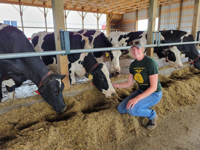 Emily Mullen, wearing a green shirt and blue jeans, kneels next to her dairy cows in a free stall barn for a photo.