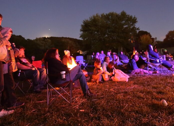 people sit in lawn chairs and on the ground, bathed in light from campfires